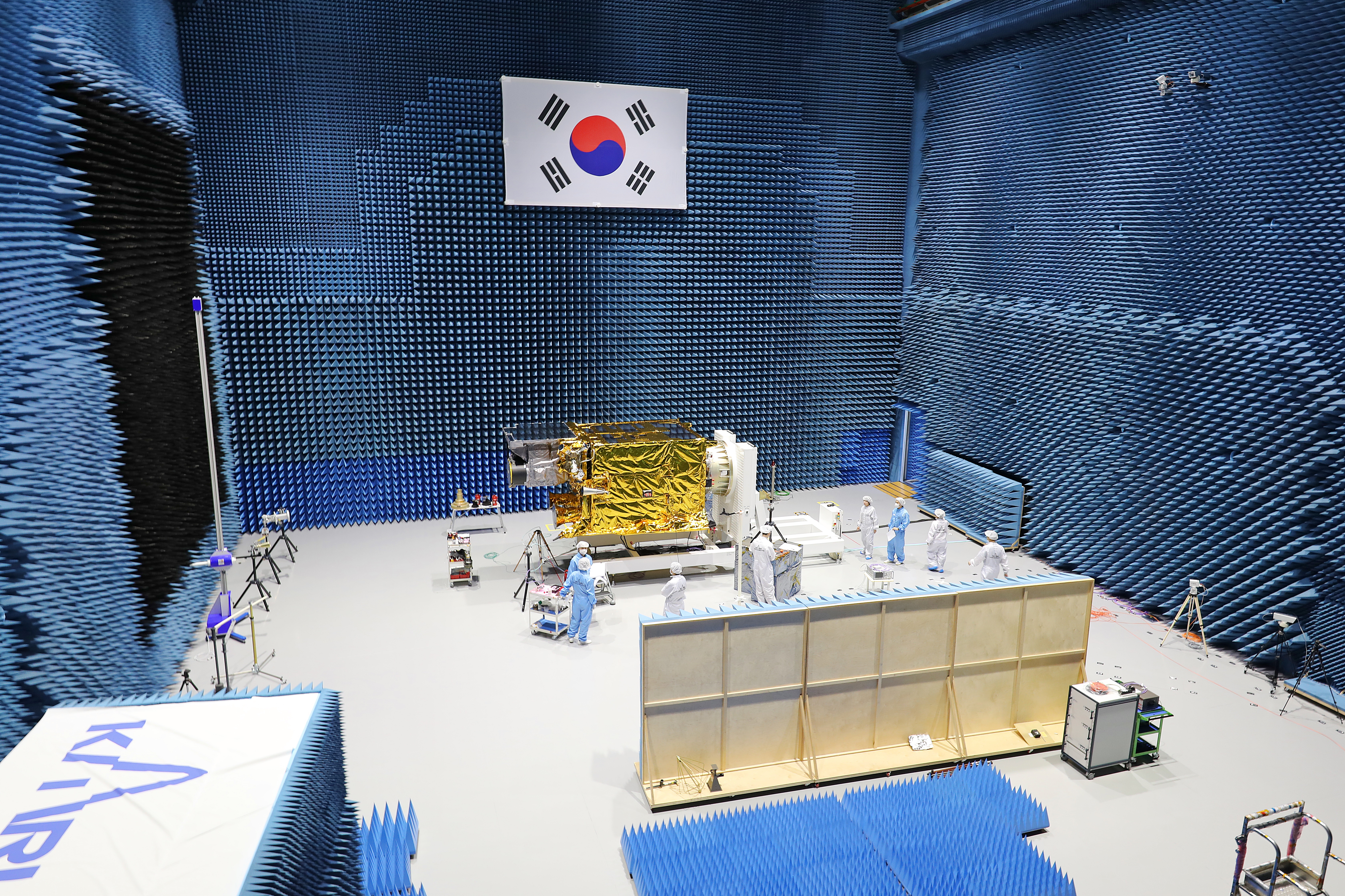 전자파 흡수체로 둘러싸인 대형 EMC(전자파 적합성) 시험실 내부에서 금색 방열재로 둘러싸인 위성이 흰색 받침대에 놓여 있고, 방진복을 입은 여러 연구원들이 주변에서 장비를 조작하며 점검하고 있는 모습. 벽면에는 태극기가 걸려 있음.