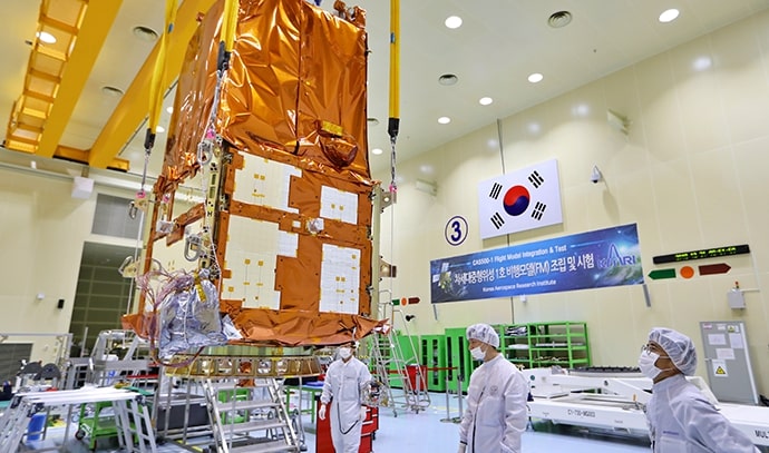 태극기가 걸린 청정실에서 크레인에 매달린 금색 방열재로 둘러싸인 위성이 공중에 떠 있는 모습. 방진복을 입은 연구원들이 아래에서 위치를 확인 중.
