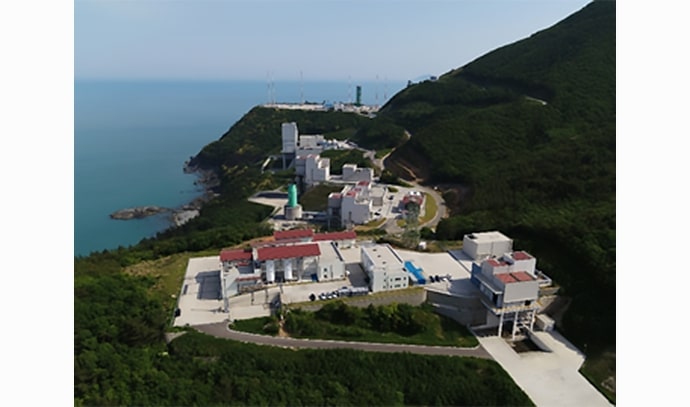 해안과 접한 나로우주센터 전경을 항공 시점에서 촬영한 이미지로, 다수의 시험설비 건물들이 초록 산지 사이에 자리 잡고 있다.